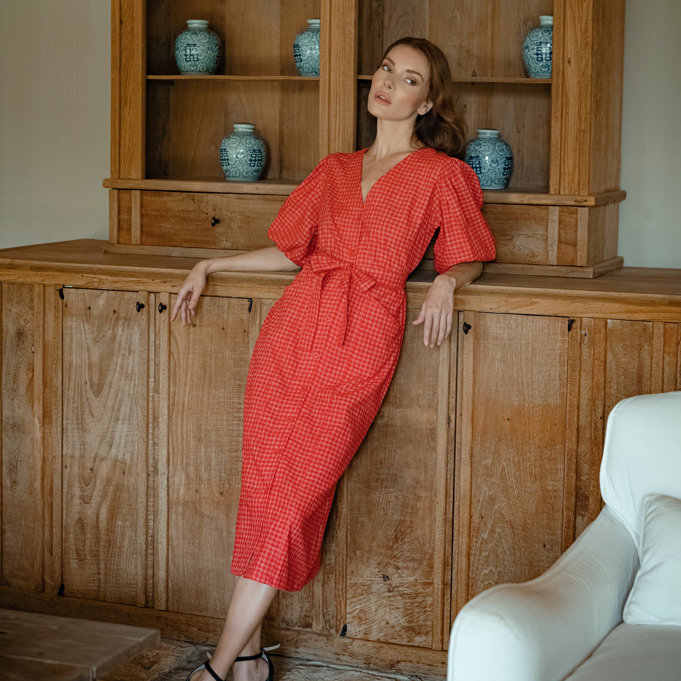 Woman in a red dress leaning against a wooden cabinet with decorative vases.