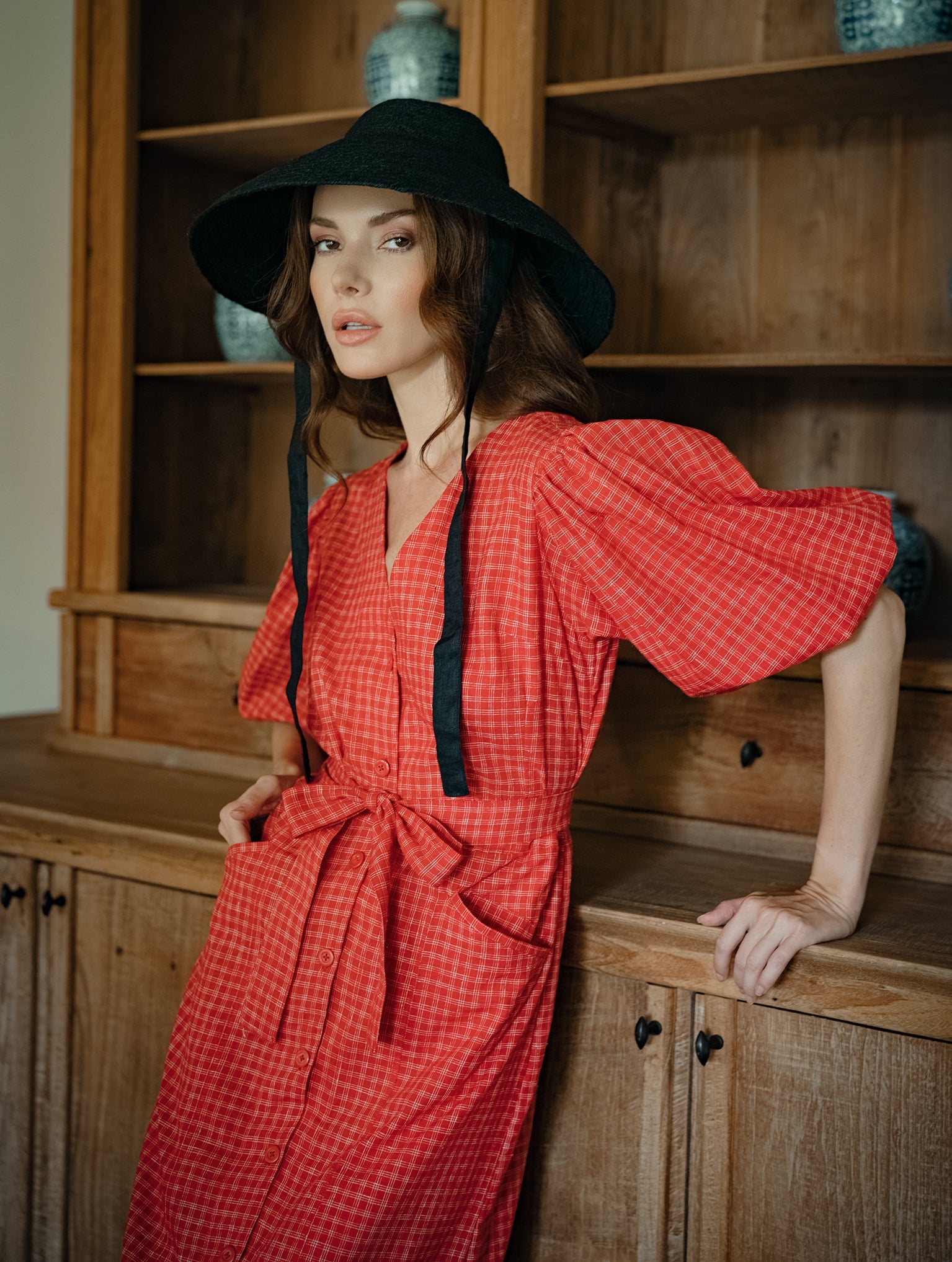 Woman in a red dress and black hat leaning against a wooden cabinet.