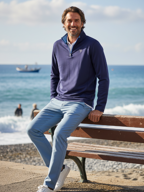 Man sitting on a bench by the ocean wearing a blue sweater and light jeans.