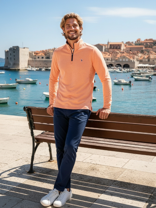 Man in peach long-sleeve shirt and navy pants standing on a bench by a waterfront with boats and buildings in the background.