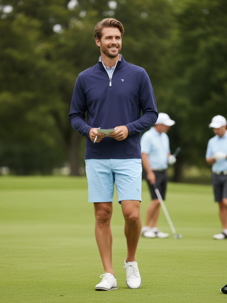 Man in navy sweater and light blue shorts standing on a golf course with trees in the background