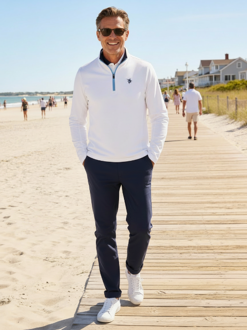 Man walking on a wooden boardwalk by the beach wearing a white sweater and navy pants.