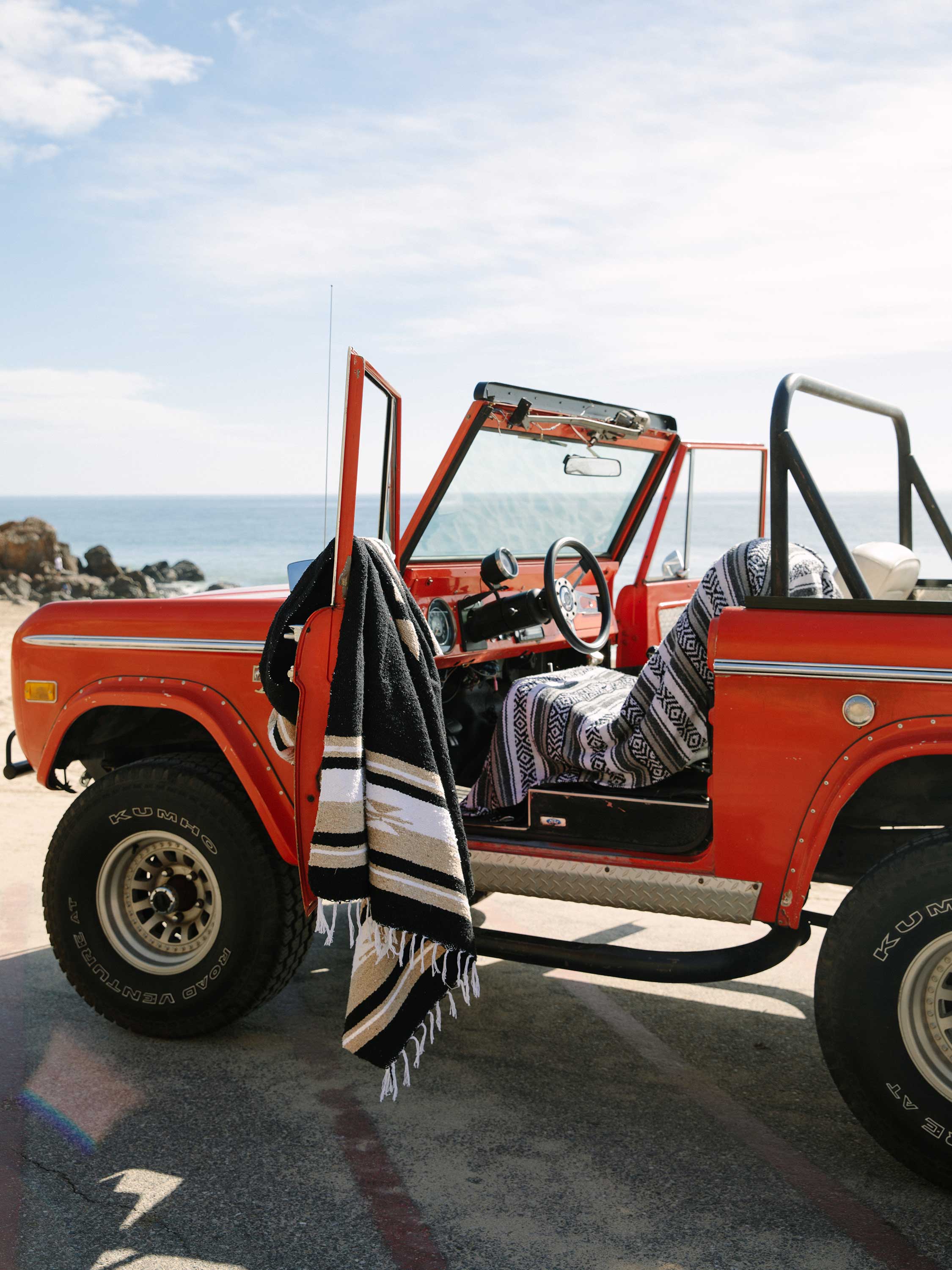 Red off-road vehicle with an open door, blanket draped over it, on a beach.