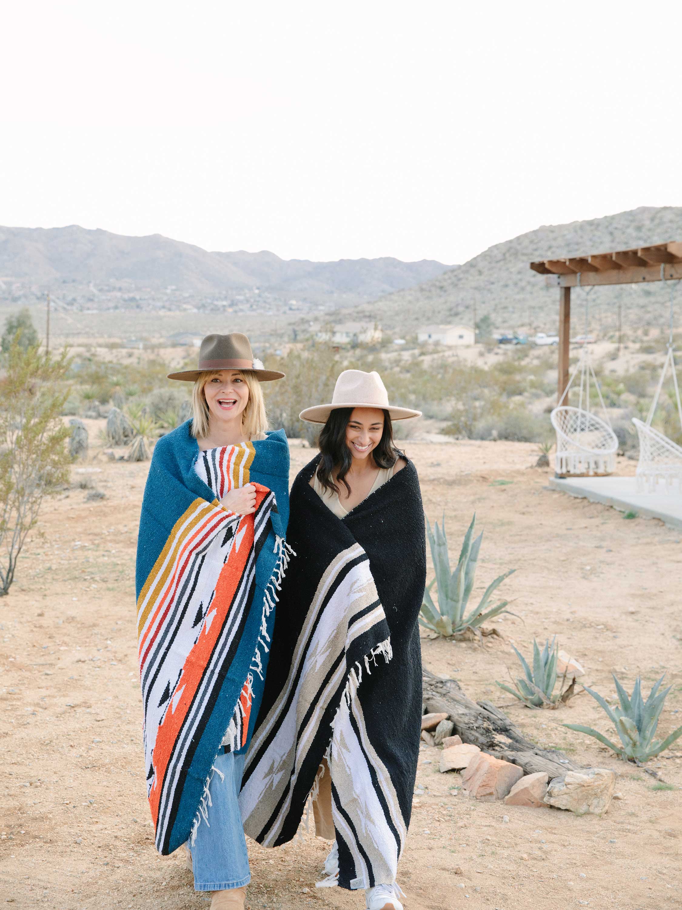 Two women standing in a desert landscape, each holding a patterned blanket.