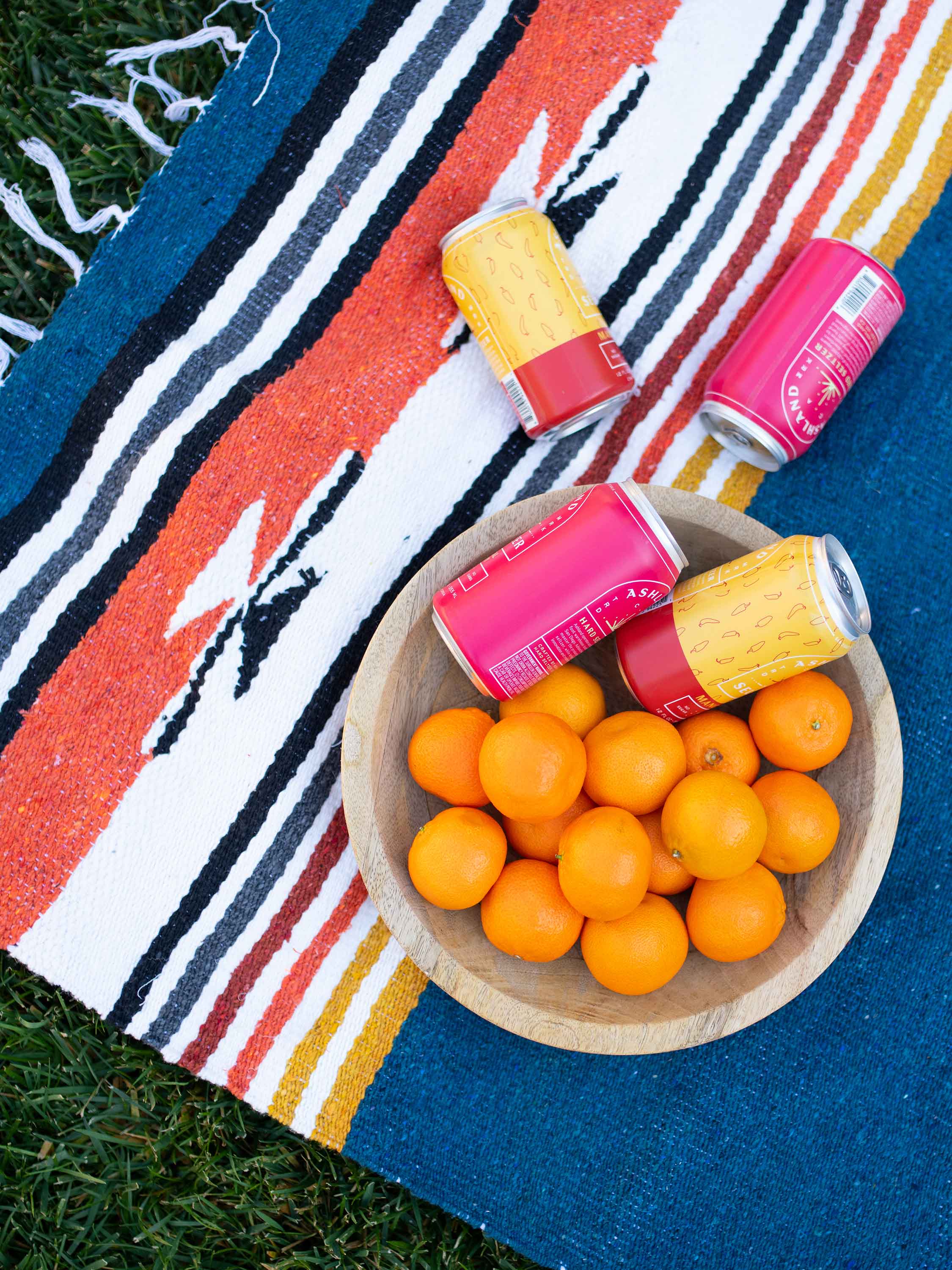 Wooden bowl with oranges and pink drinks on a colorful striped blanket outdoors