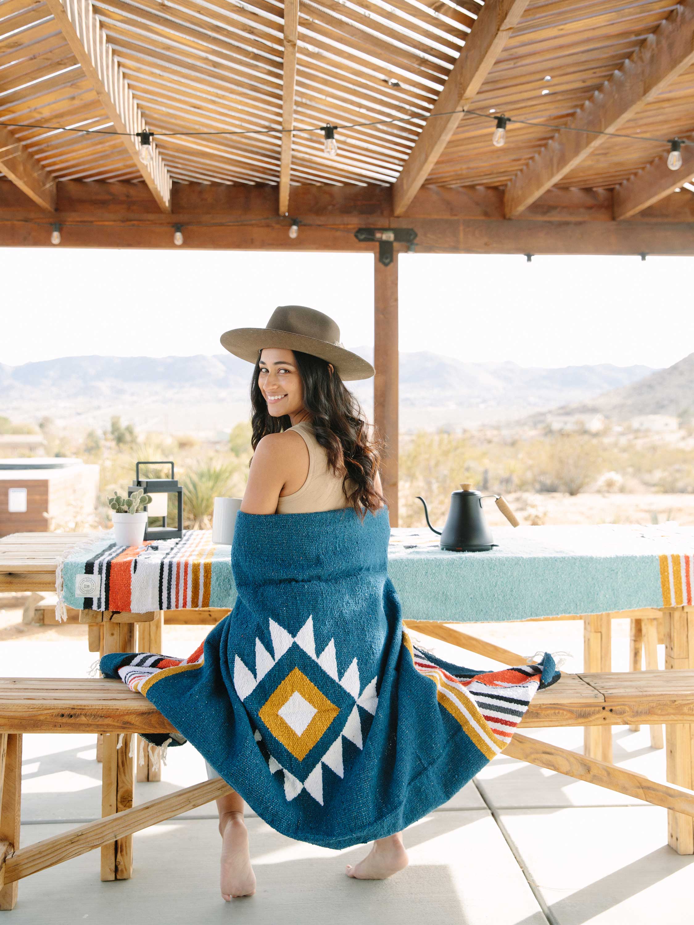 Woman sitting on a wooden bench with a blue patterned blanket, wearing a hat, in an outdoor setting.