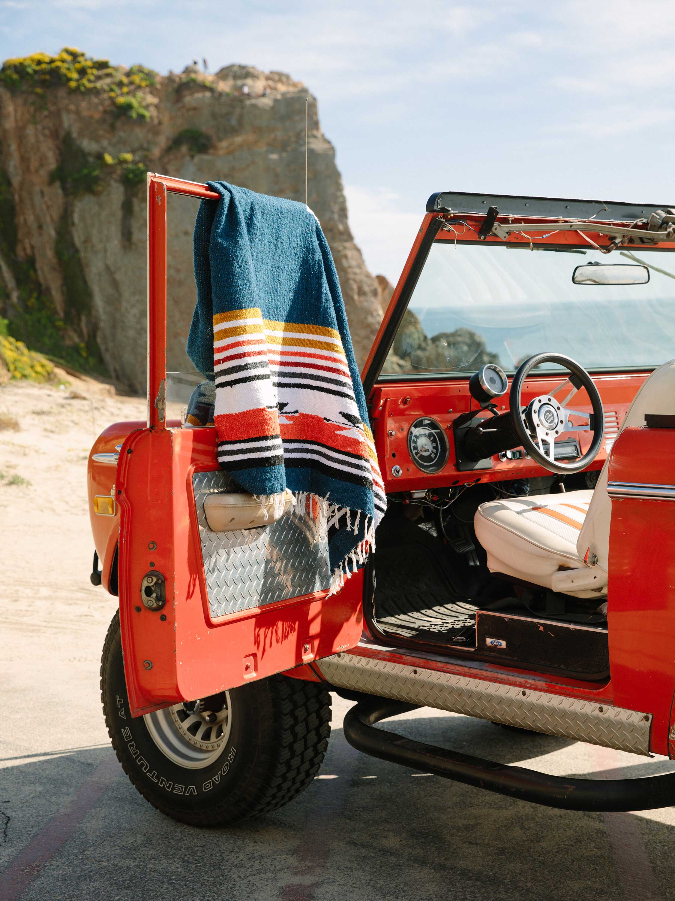 Red vintage jeep with a colorful towel draped over the door, parked on a scenic road.