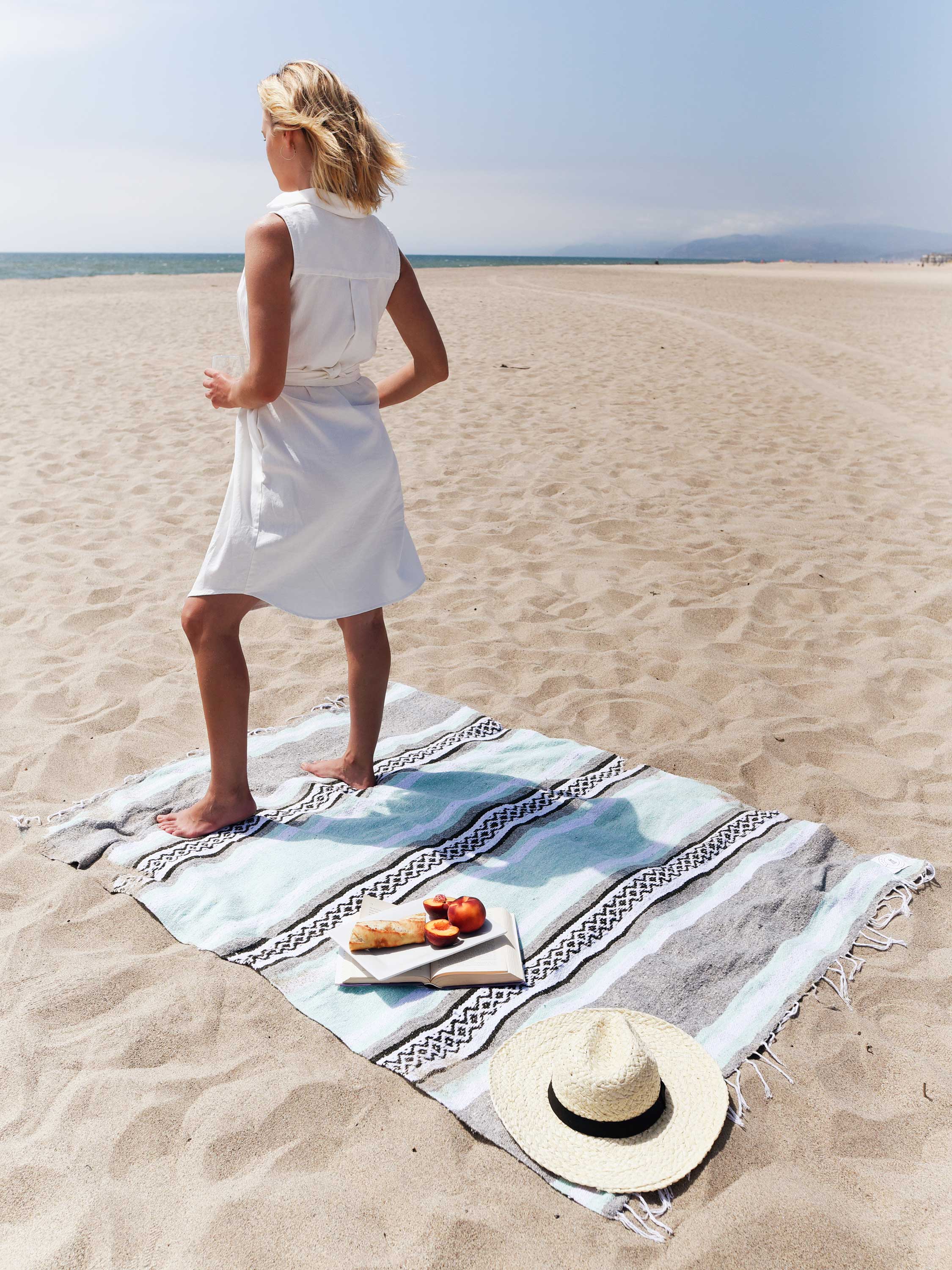 Woman in a white dress standing on a beach with a picnic setup.