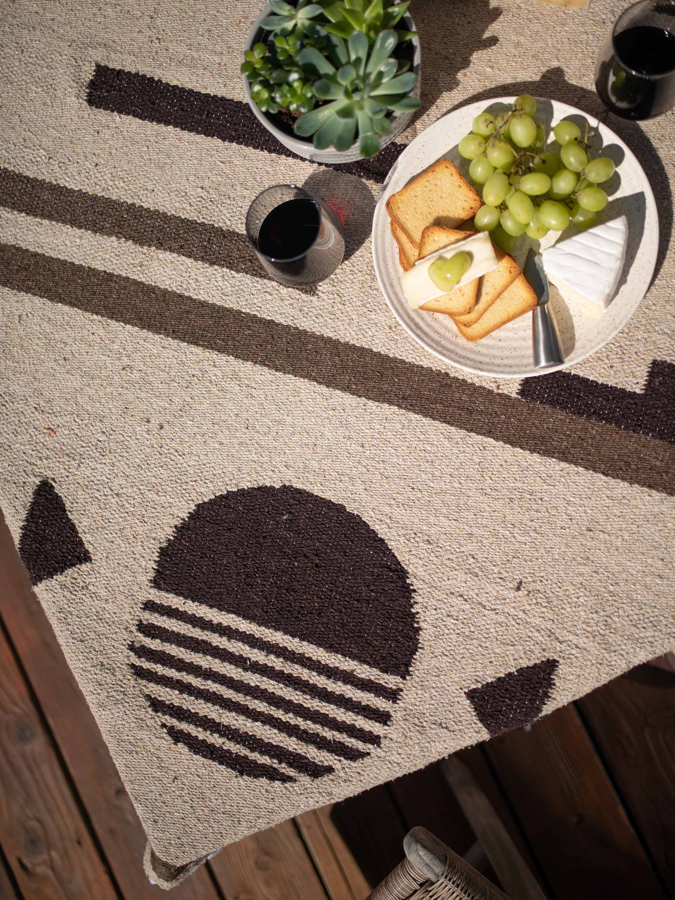 Outdoor setting with a patterned rug, plate of food, and glasses on a wooden surface.