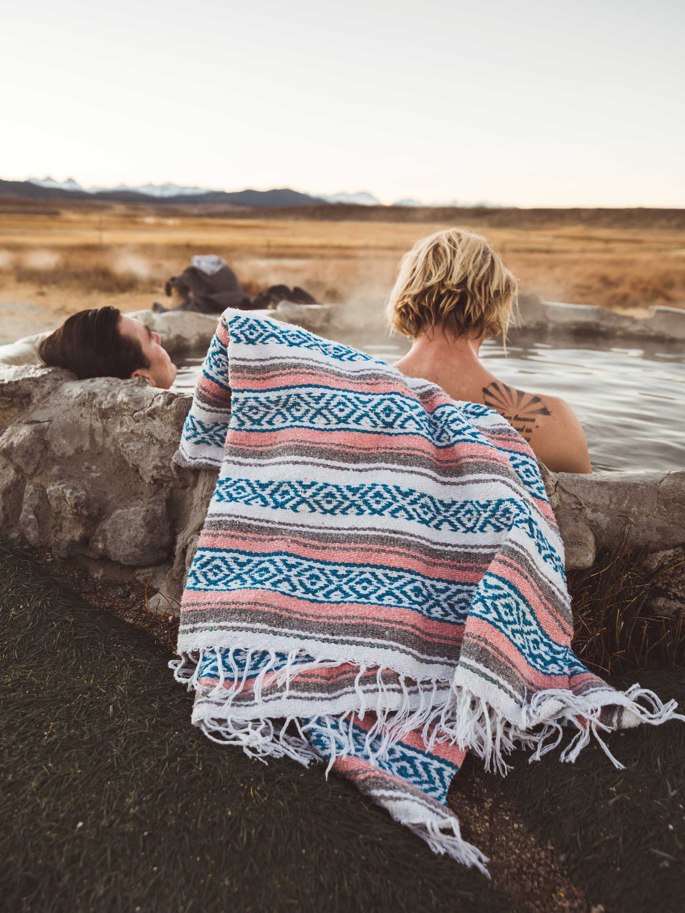Two people relaxing in a hot spring with a colorful patterned blanket.