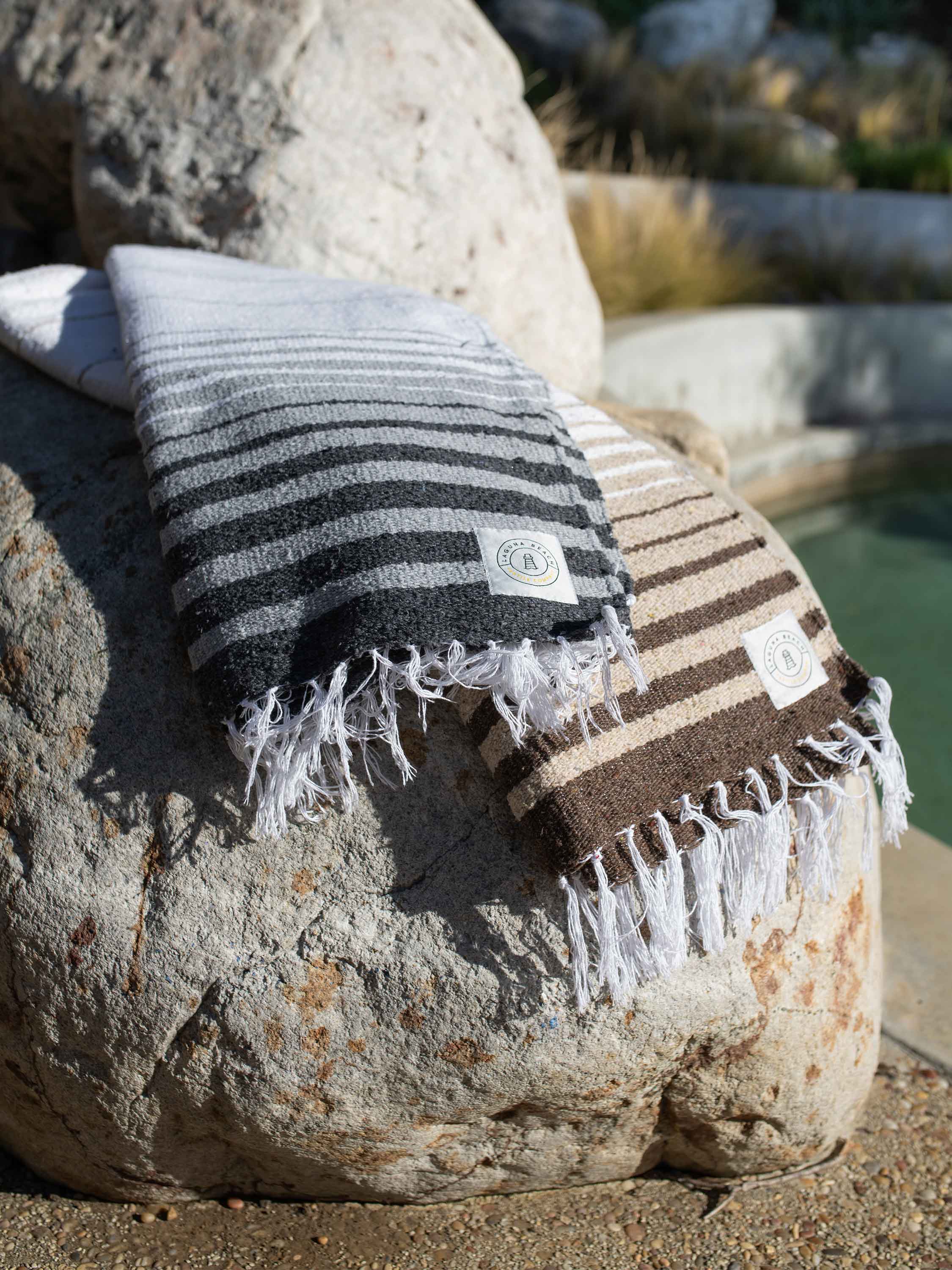 Two striped towels, one black and white and one brown and white, draped over a rock by a body of water.