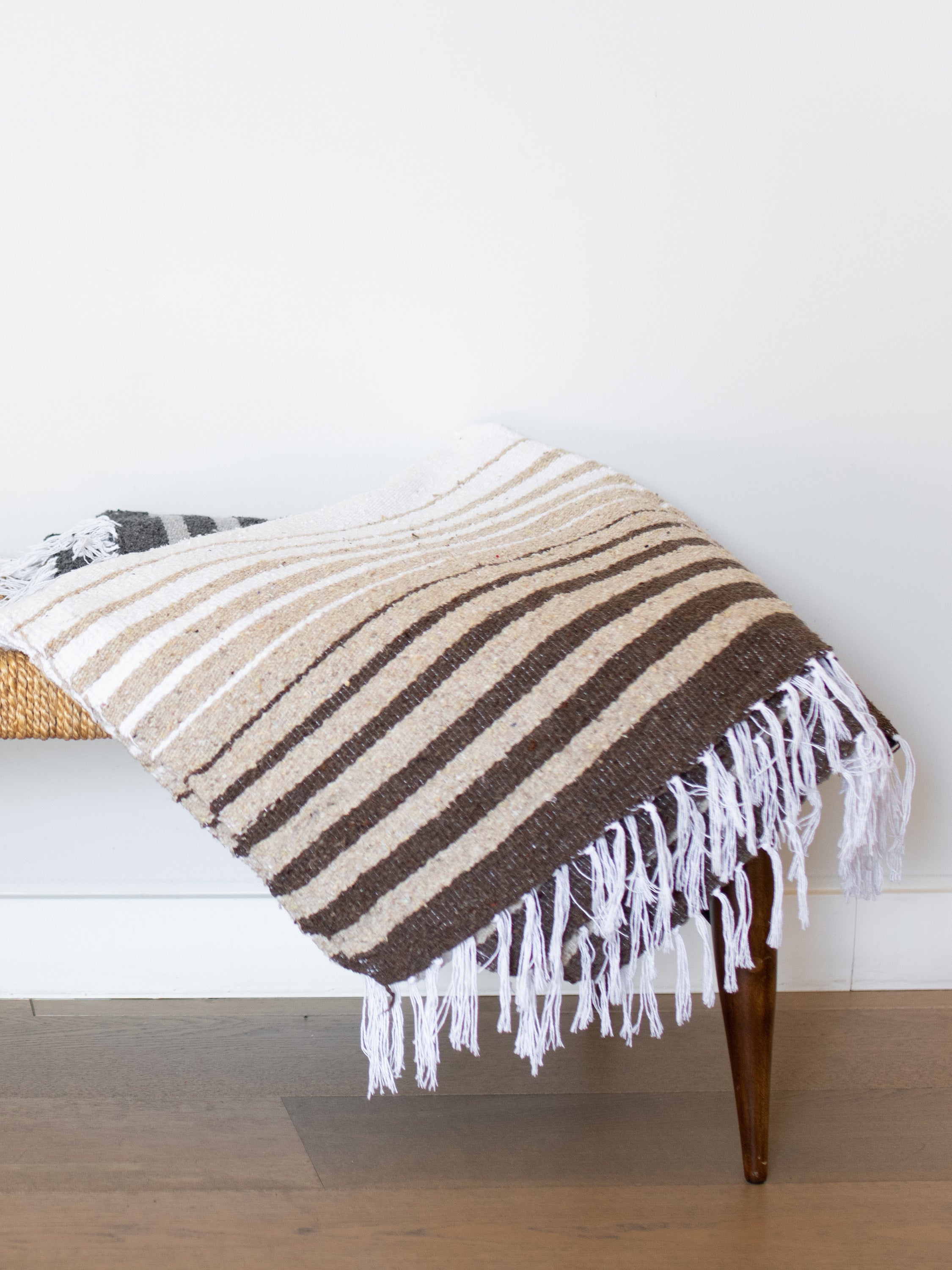 Striped blanket with fringes draped over a wooden bench against a white wall.