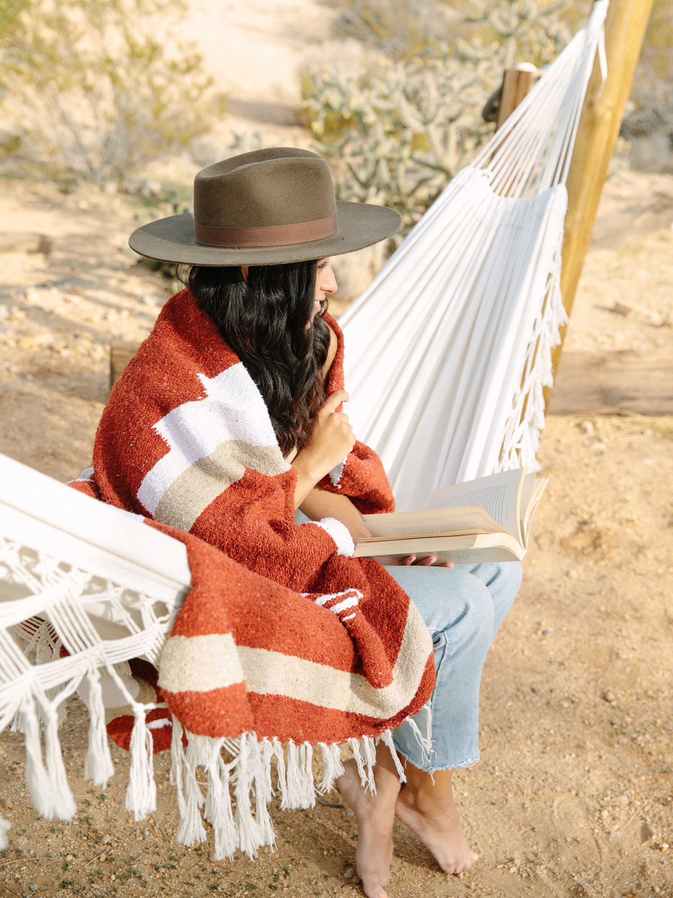 Person sitting in a hammock wearing a red blanket and hat, surrounded by natural elements.