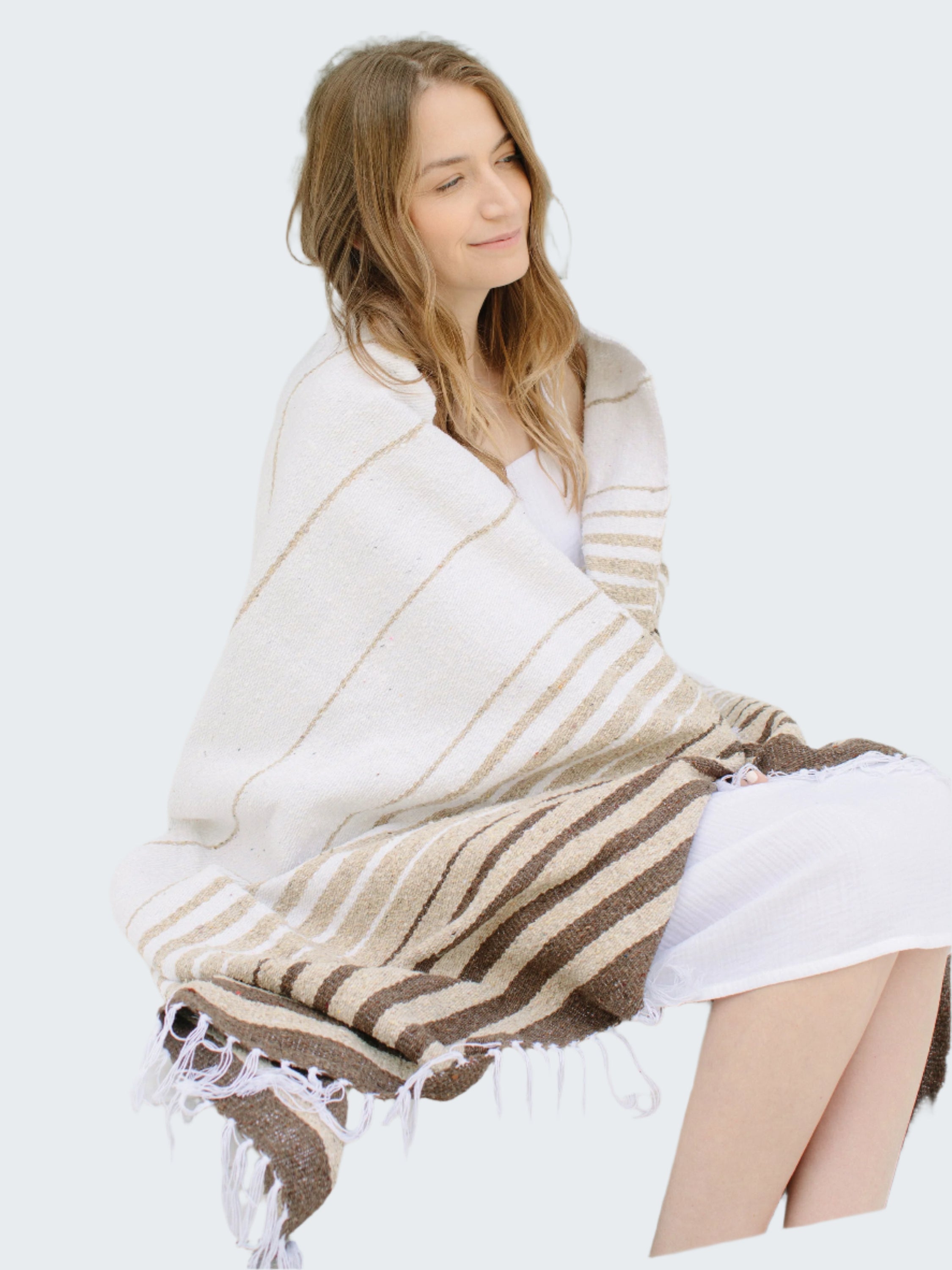 Woman sitting on a bench wrapped in a striped blanket with a neutral background