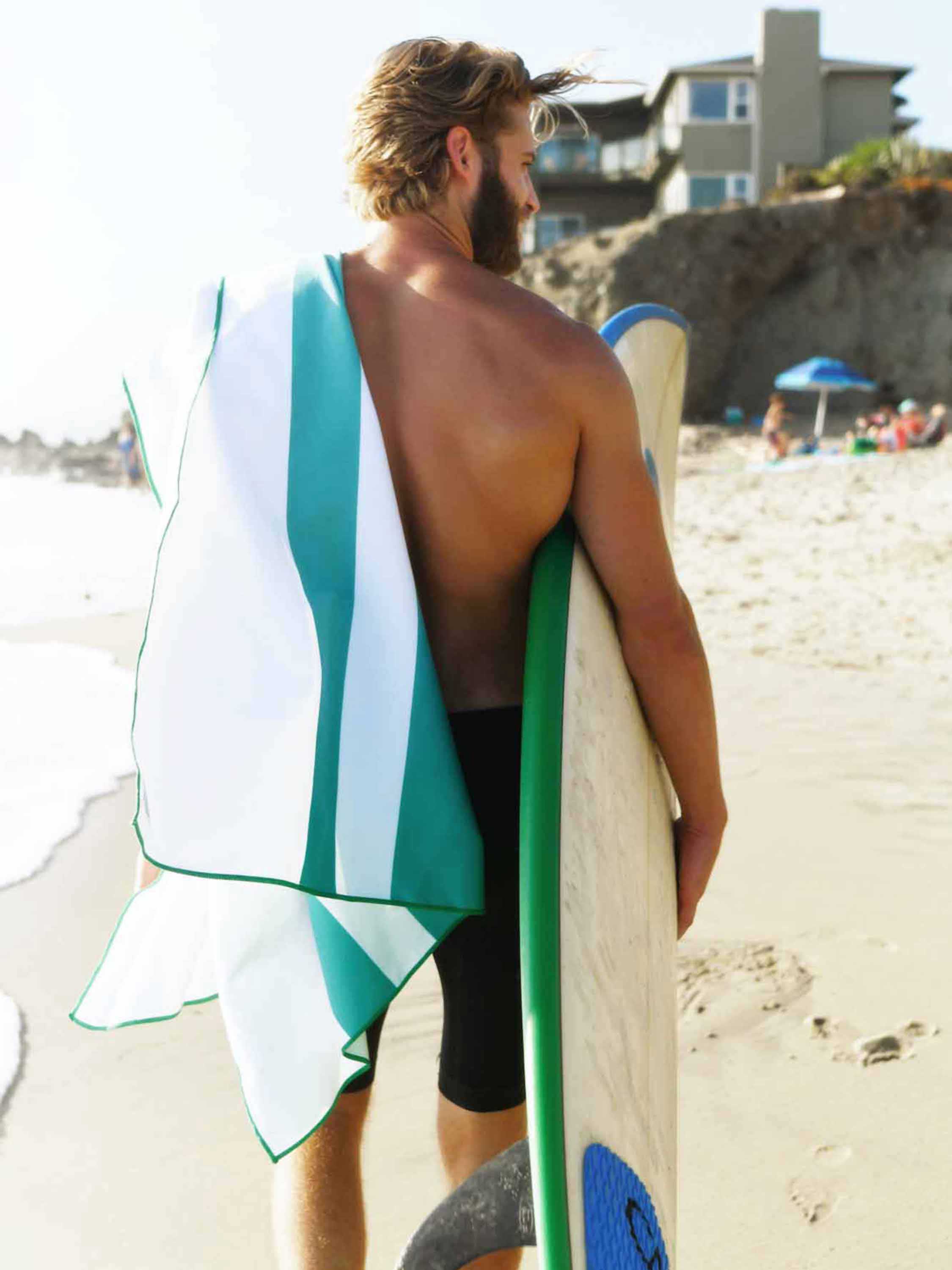 Man holding a surfboard with a towel draped over his shoulder on a beach.