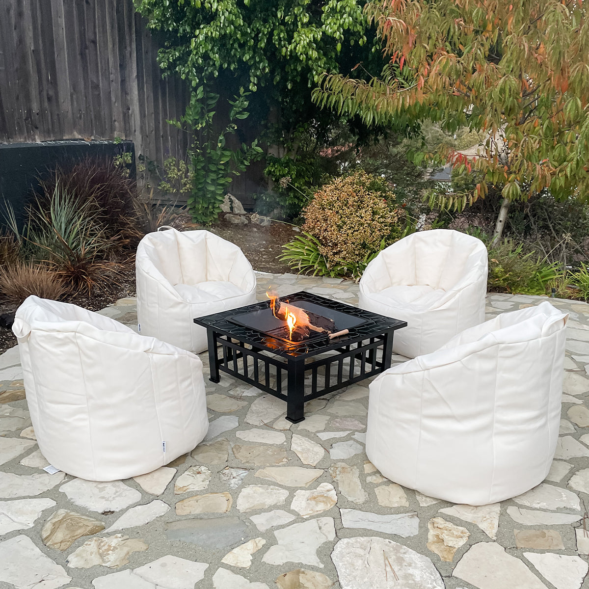 White bean bag chairs around a fire pit on a stone patio with greenery in the background