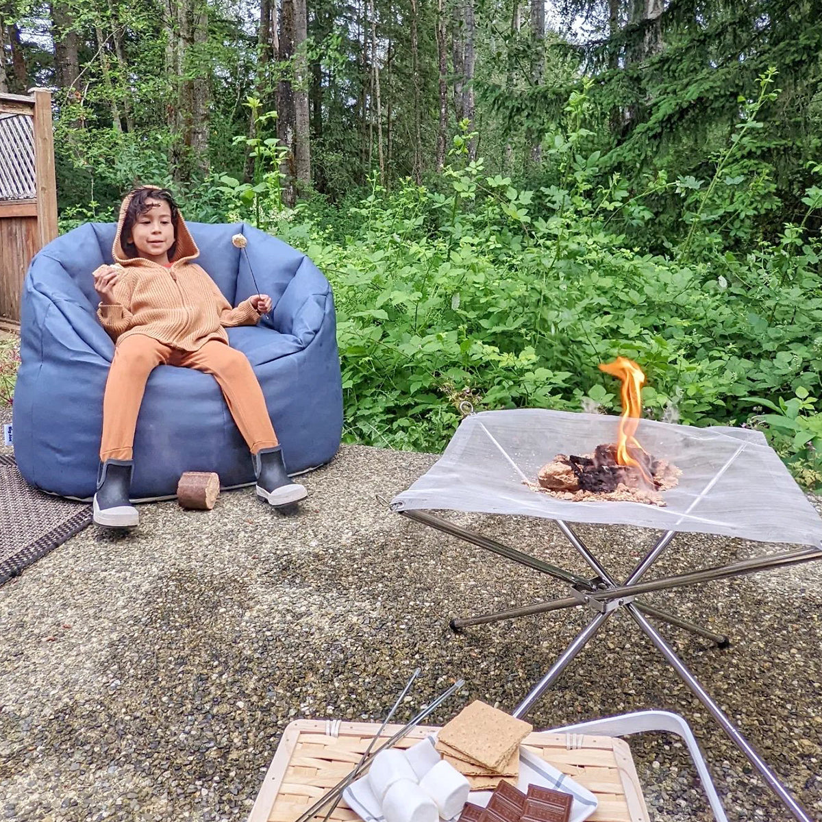 Child sitting on a blue bean bag chair by a campfire in a forest setting