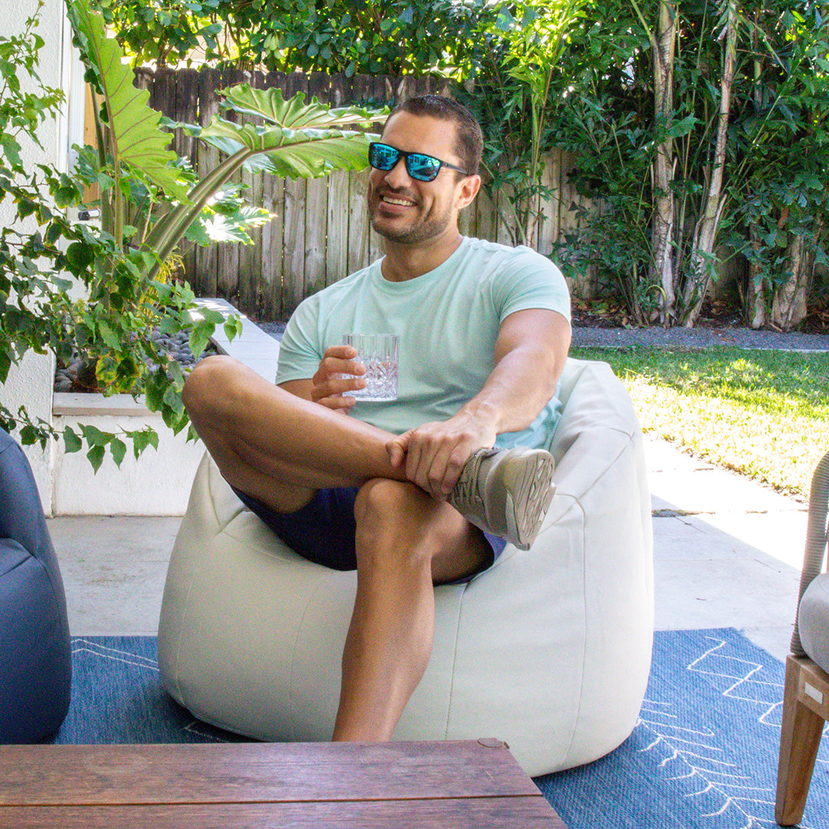 Man sitting on a bean bag chair outdoors with plants and a wooden fence in the background