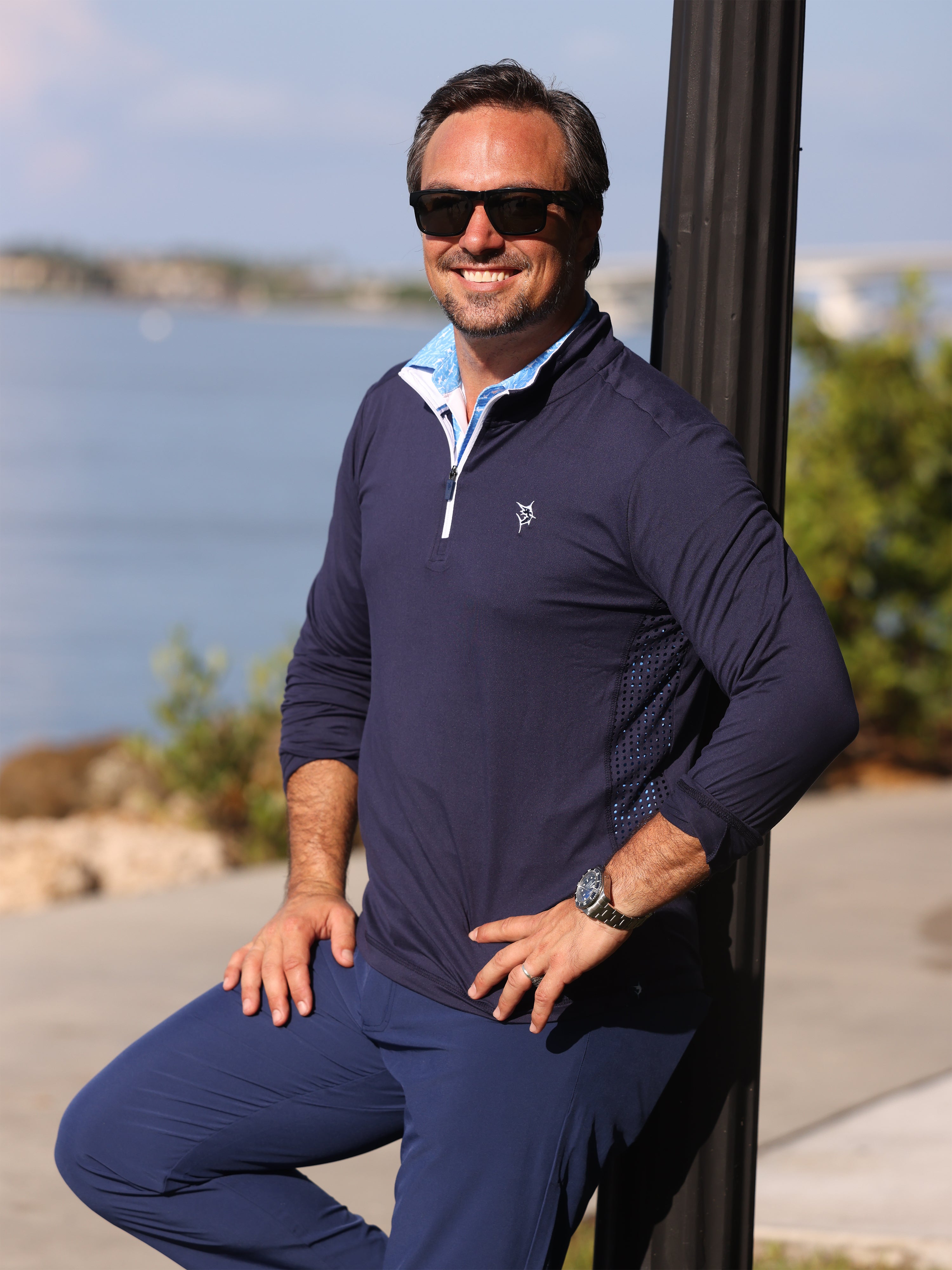 Man wearing a navy blue outfit with sunglasses, standing by a waterfront.