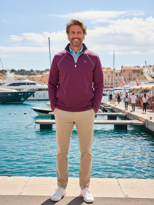 Man in a maroon pullover and beige pants standing by a marina with boats and people in the background.