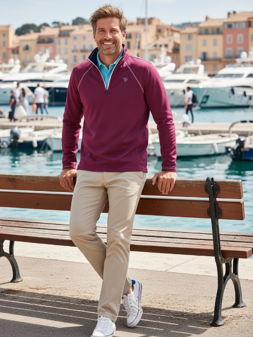 Man in a purple jacket and beige pants leaning on a bench by a waterfront with boats and buildings in the background.