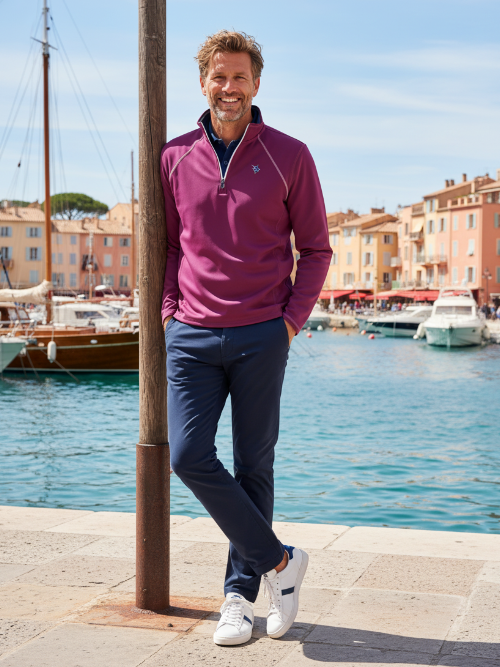 Man in a pink sweater and blue pants standing by a waterfront with boats and buildings in the background.
