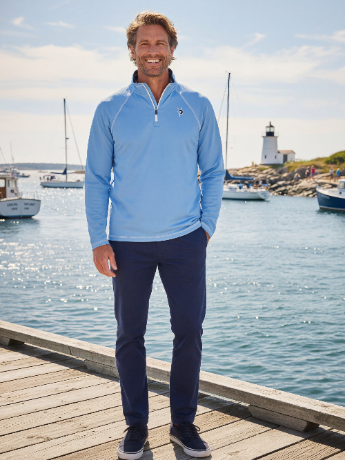 Man wearing a blue pullover with a zipper, standing on a dock by the water with boats and a lighthouse in the background.