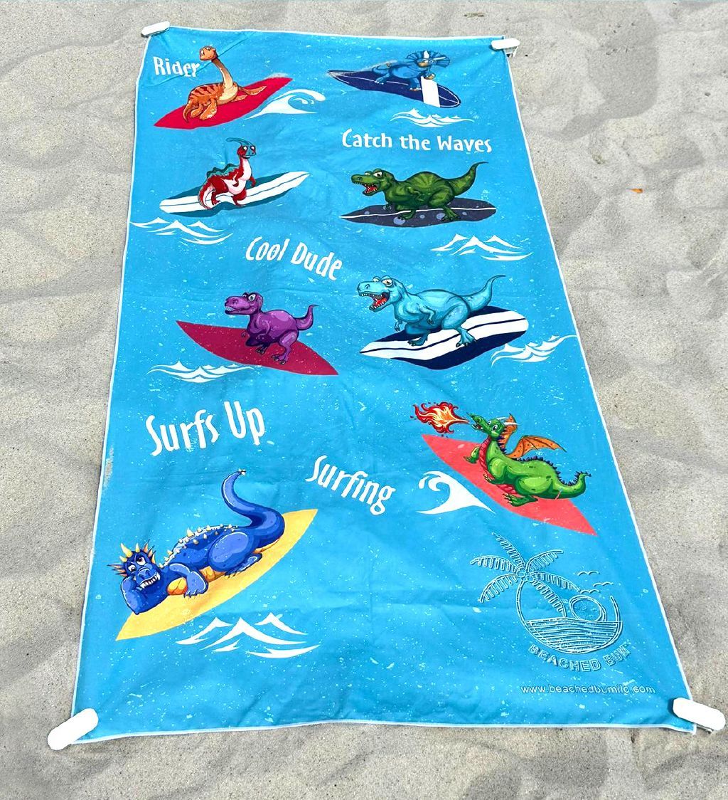 Beach towel with cartoon dragons surfing on a sandy background