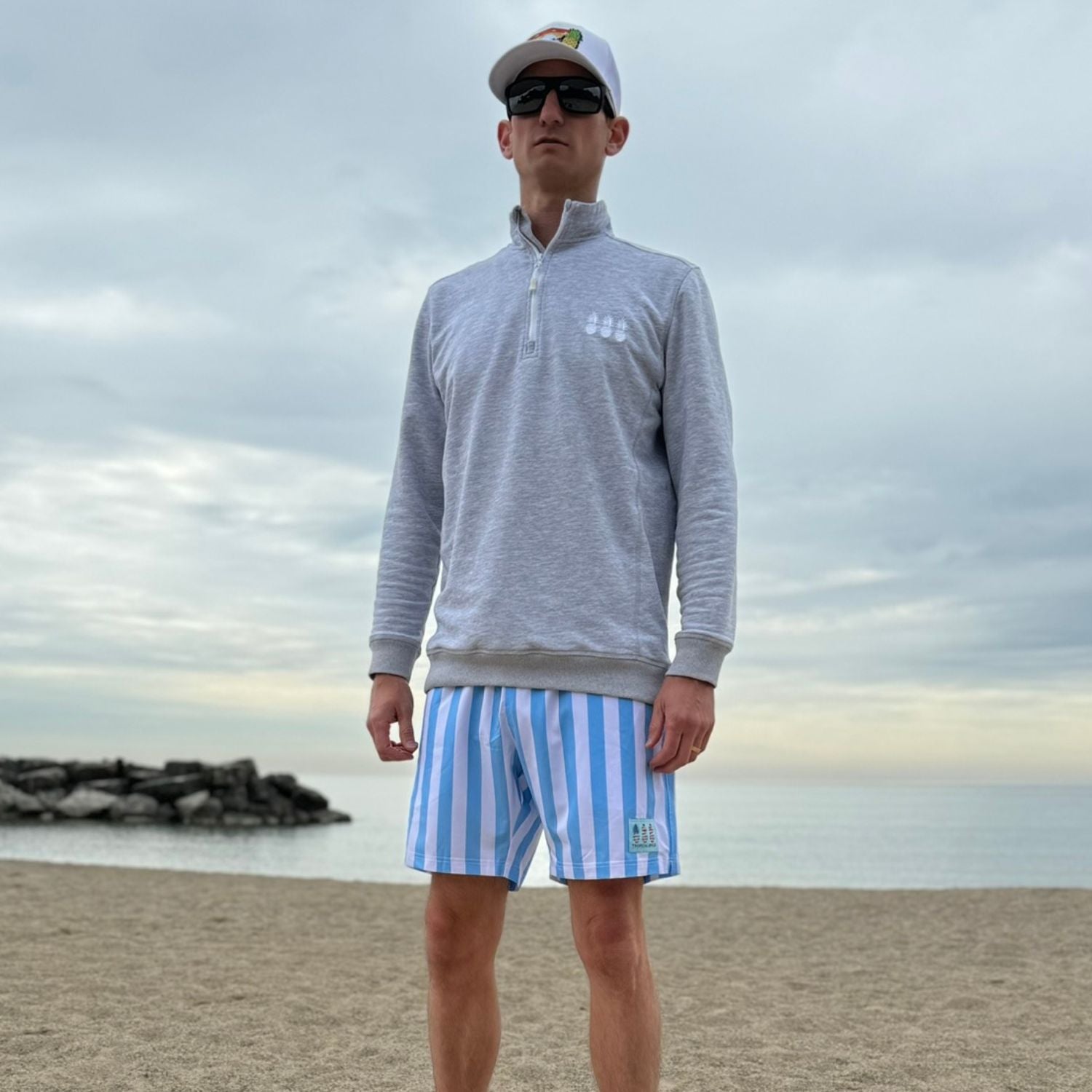 Man wearing a gray sweater and blue striped shorts standing on a beach with a cloudy sky.