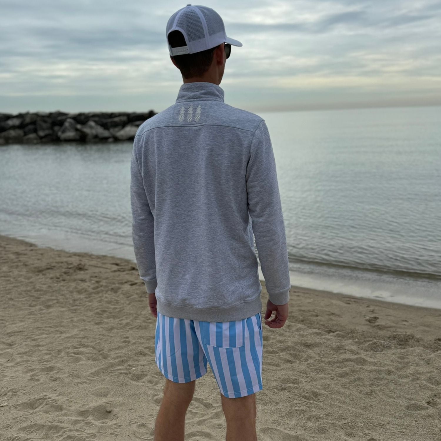 Person wearing a gray jacket and blue striped shorts standing on a beach.