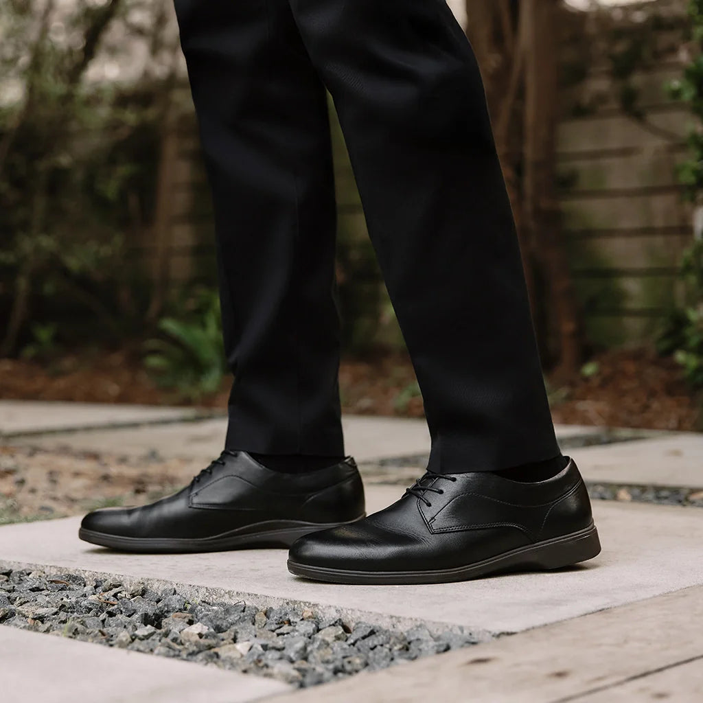 Person wearing black dress shoes and pants standing on a concrete surface with a blurred natural background