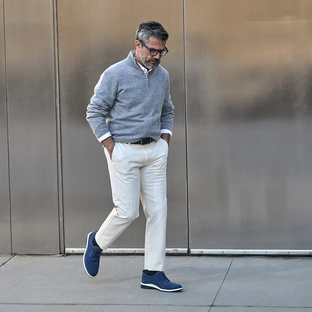 Man in gray sweater and white pants walking against a metallic wall.