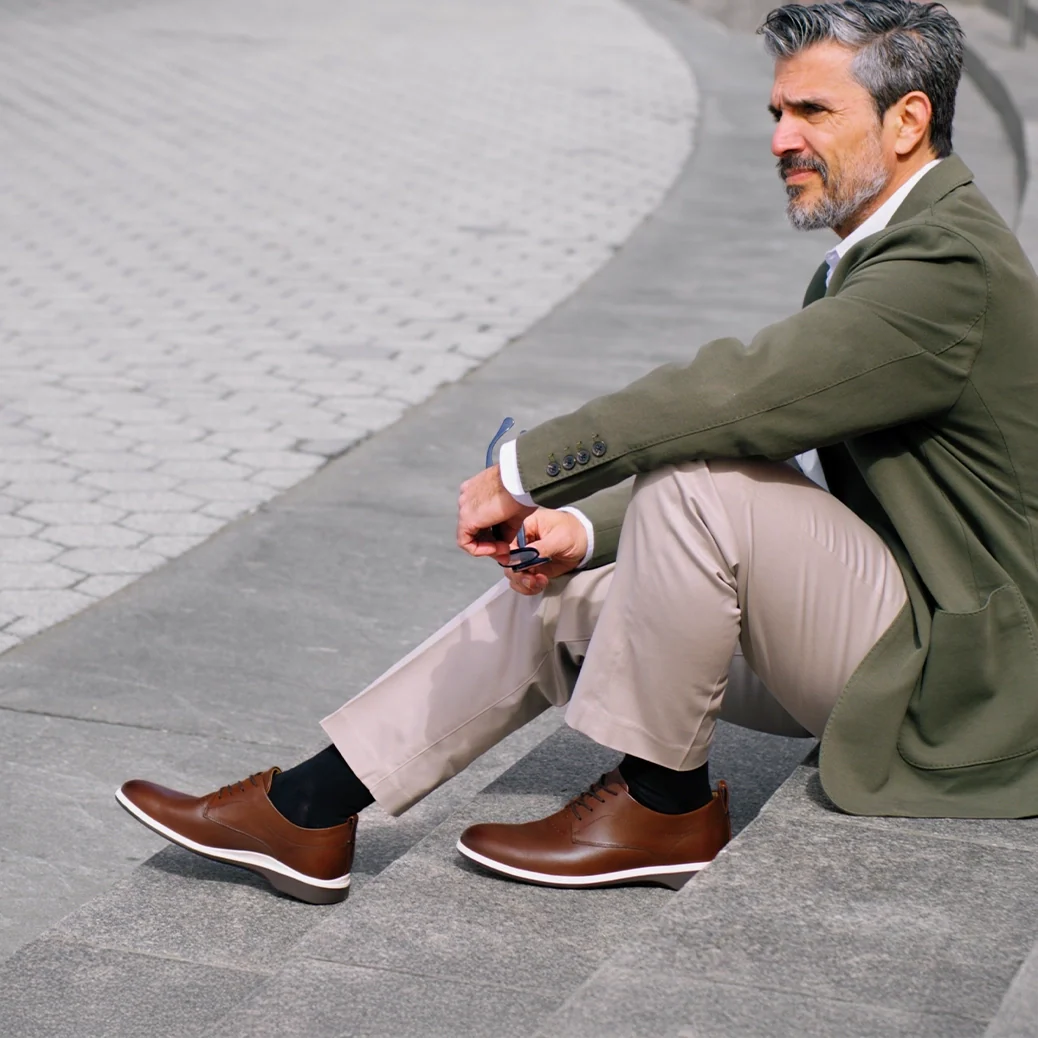 Man sitting on a stone bench wearing brown shoes, green coat, and beige pants.