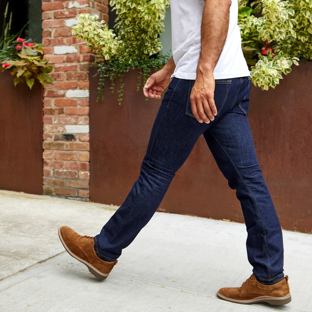 Person wearing blue jeans and brown shoes walking on a sidewalk with plants in the background