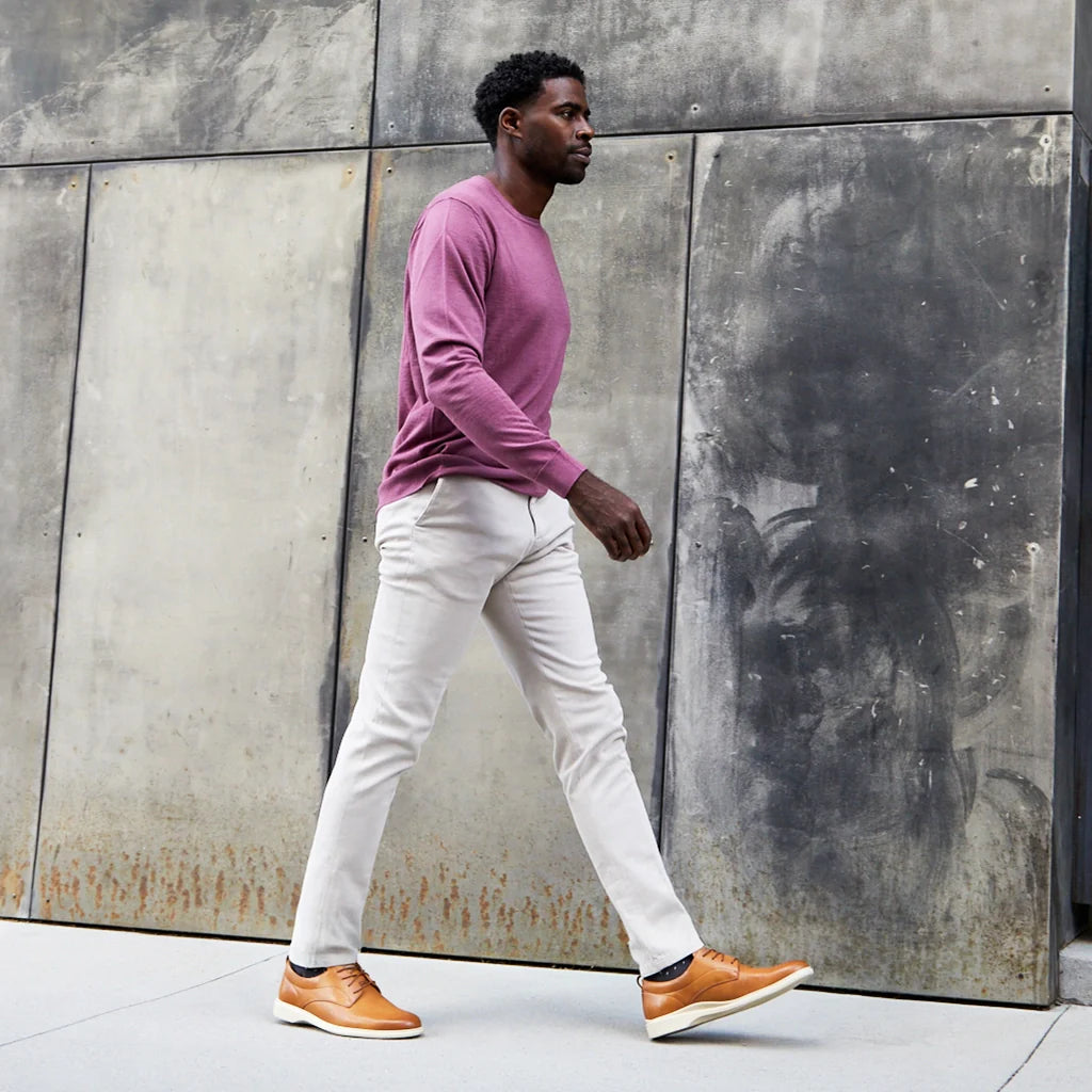 Man walking against a textured wall wearing a pink sweater, white pants, and brown shoes.