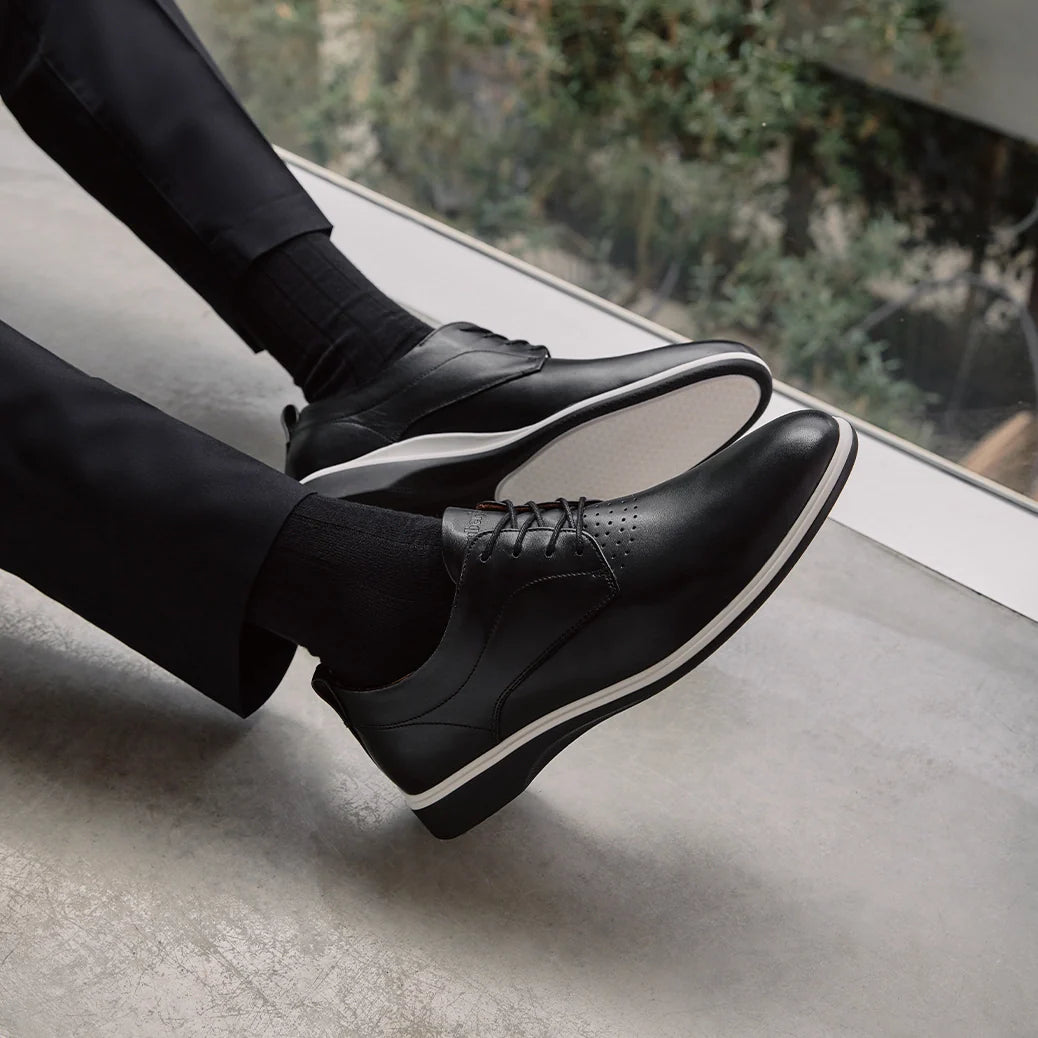 Person wearing black leather shoes with white soles on a light-colored floor.
