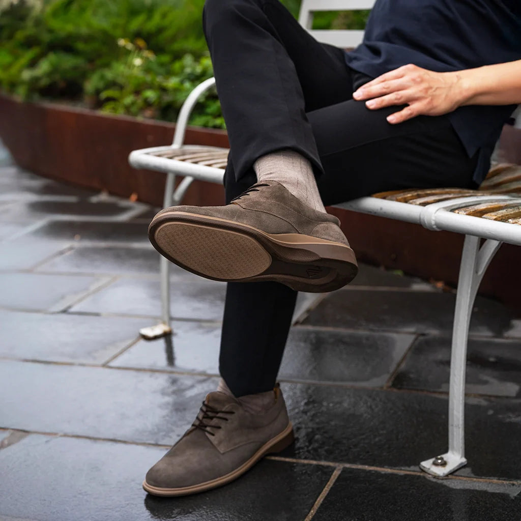 Person wearing brown shoes and black pants sitting on a bench outdoors.