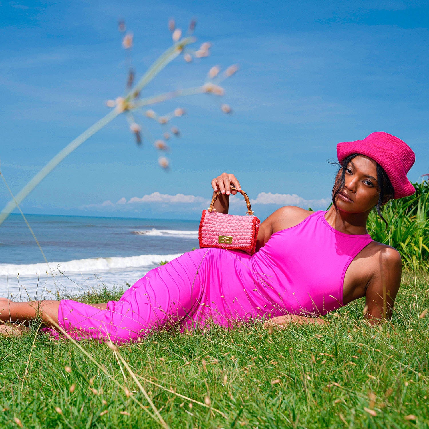 Woman in a pink dress and hat holding a pink handbag, lying on grass with ocean in the background