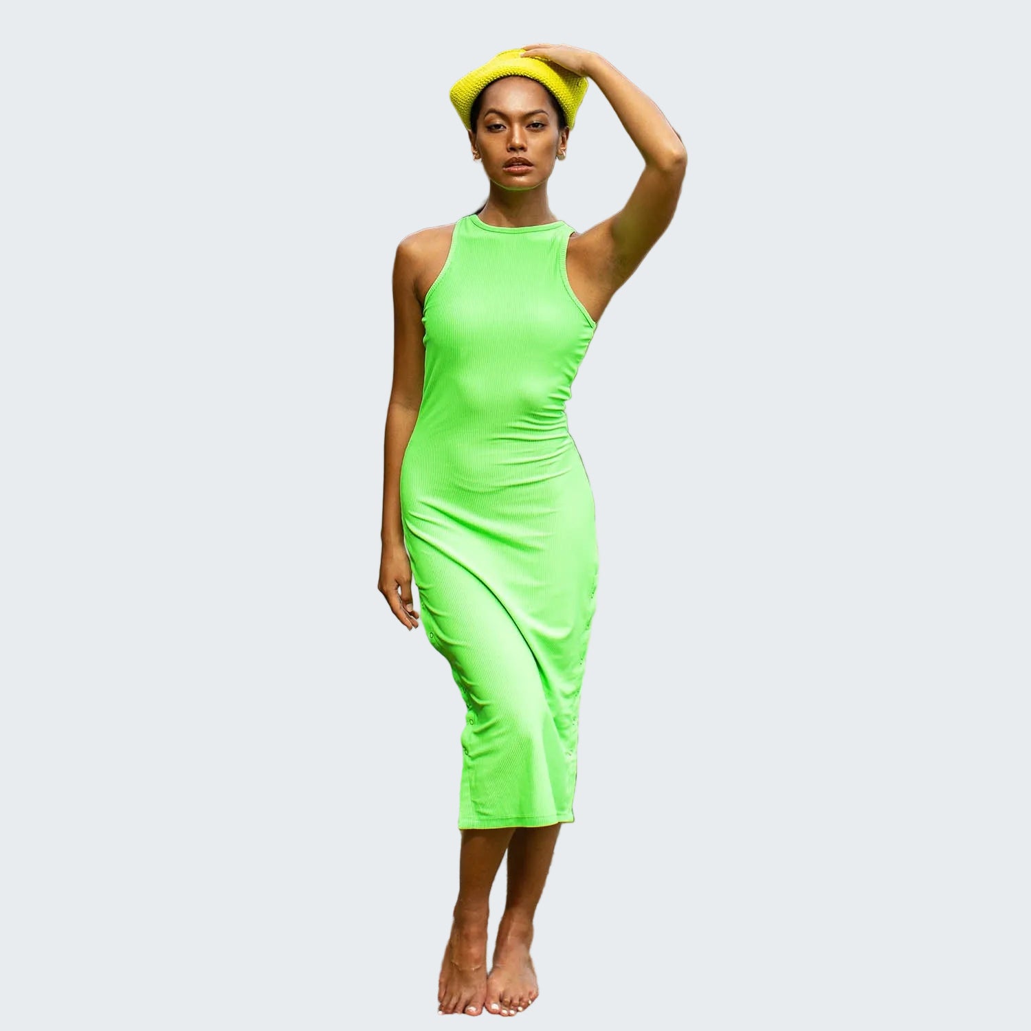 Woman in a bright green dress and yellow hat standing on a rocky beach.