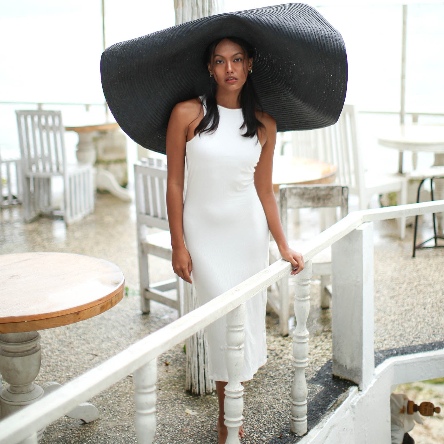 Woman wearing a large black hat in an outdoor setting with white furniture.
