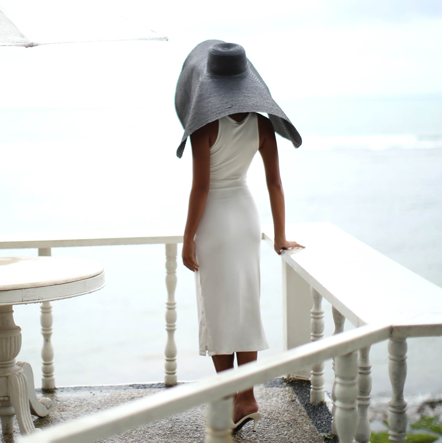 Person wearing a white dress and large gray hat on a balcony with a blurred background