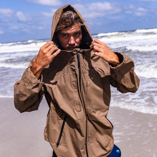 Person wearing a brown hooded jacket on a beach with waves in the background