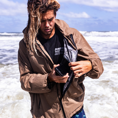 Man in a brown jacket holding a phone on a beach with ocean waves in the background