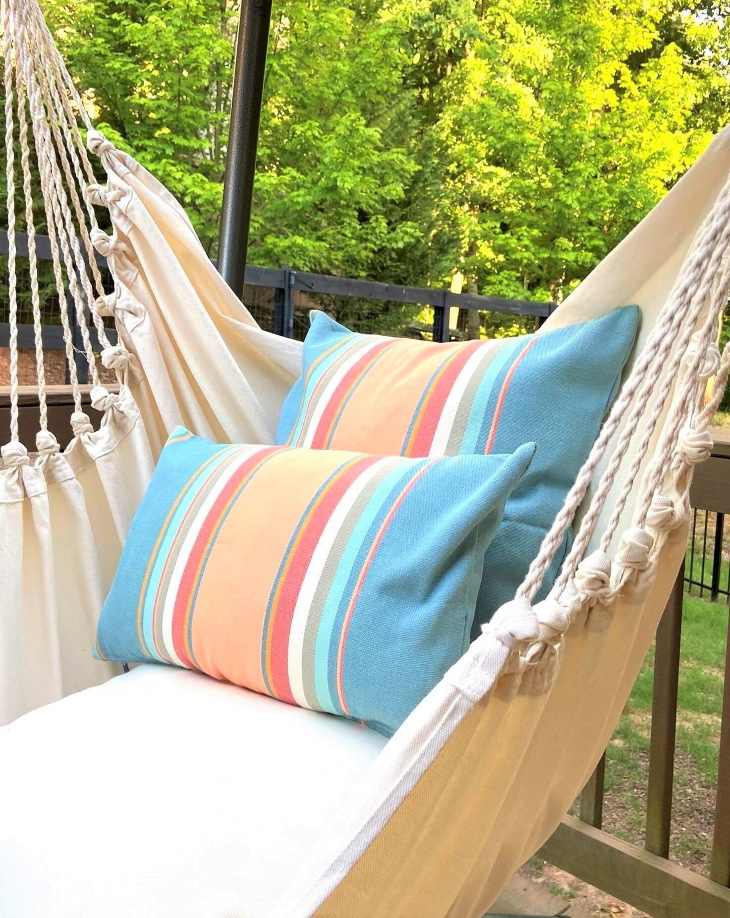 Beige hammock with colorful striped pillows on a deck with greenery in the background