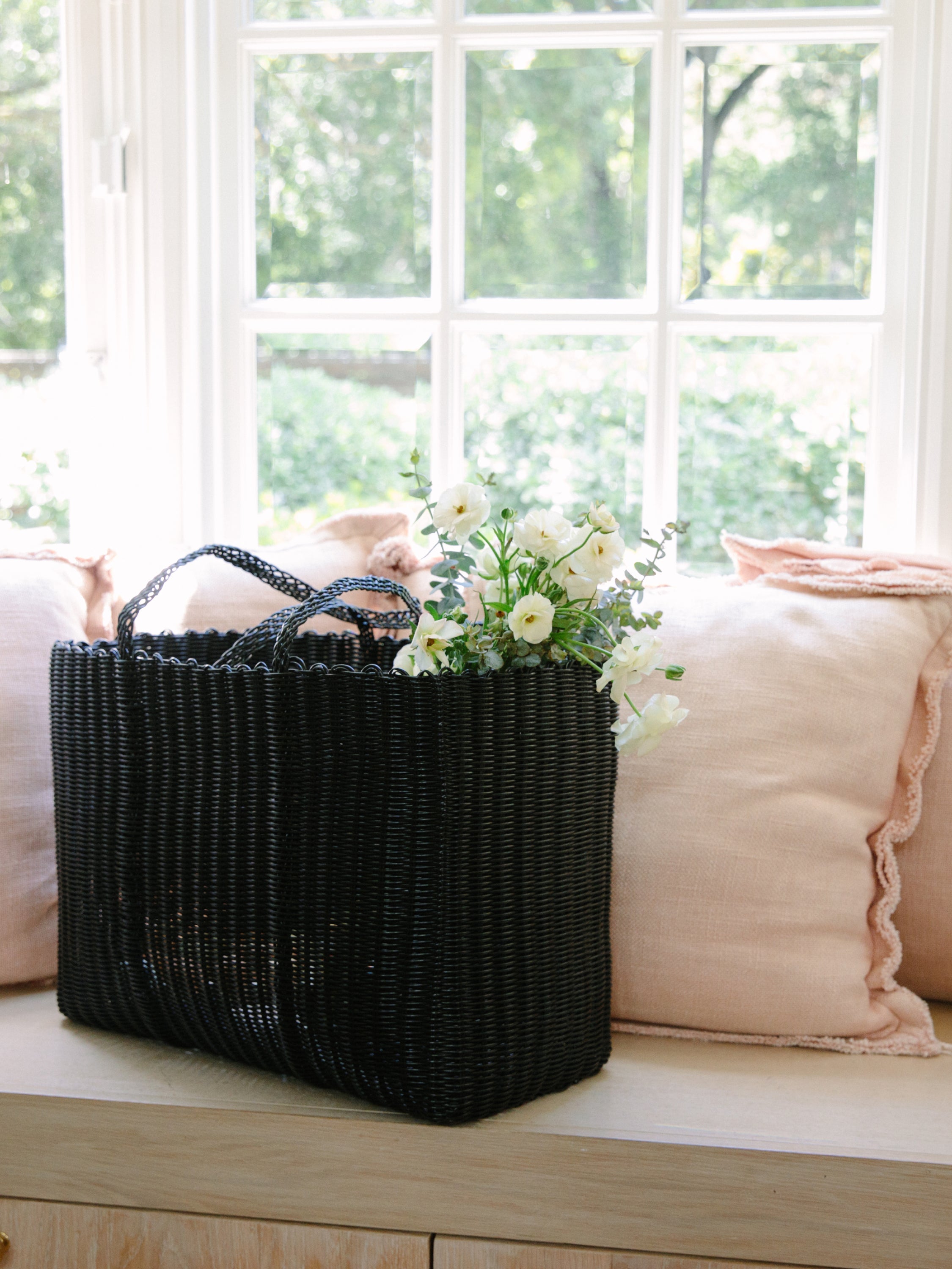 Black woven basket with flowers on a wooden bench by a window
