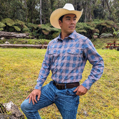 Man wearing a blue plaid shirt and cowboy hat standing in a grassy outdoor area.