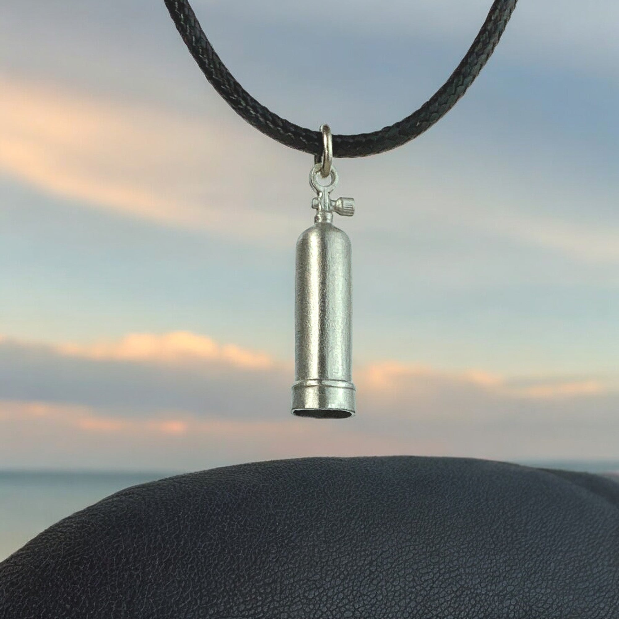Silver cylindrical pendant on a black cord against a sunset sky