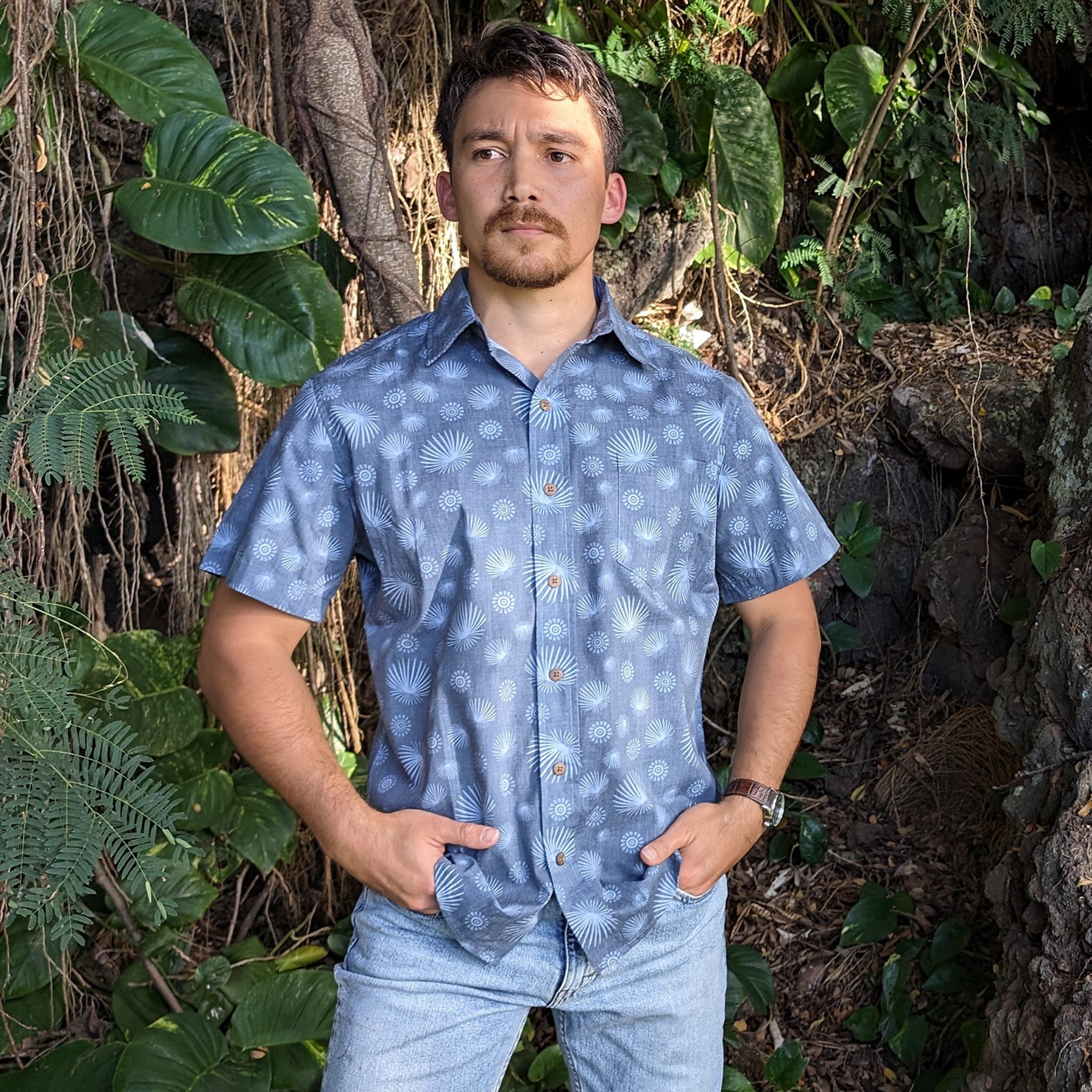 Man wearing a blue patterned shirt standing in a natural setting with greenery.