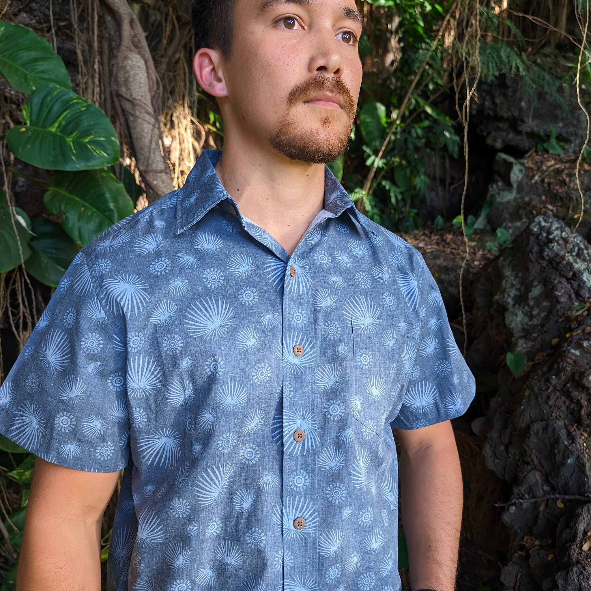 Man wearing a blue patterned shirt standing in a natural setting with greenery.