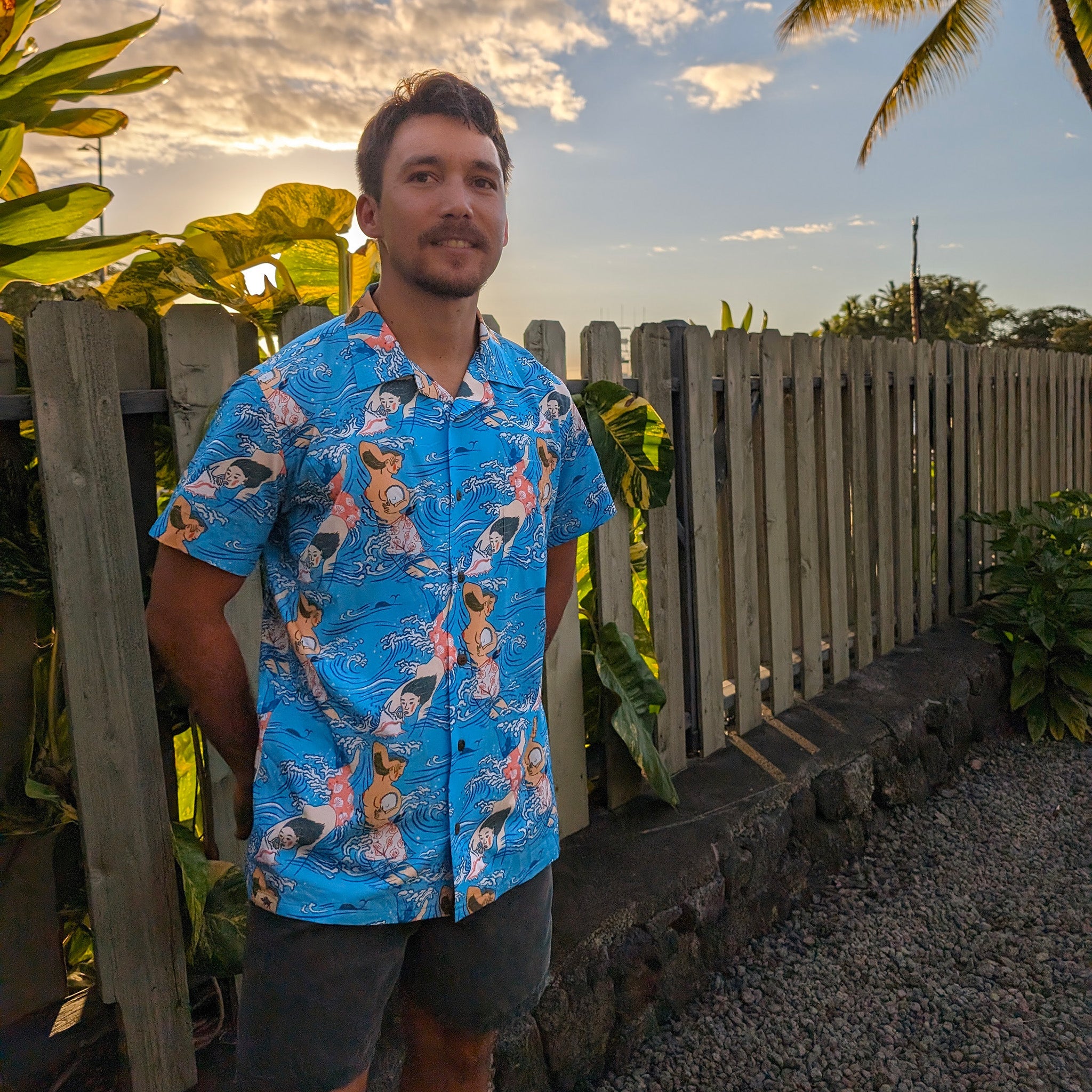Man wearing a blue patterned shirt standing outdoors with plants and a wooden fence in the background.